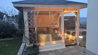 Ein gemütlicher Holzpavillon mit warmer Beleuchtung, Carport- oder Terrassenplane mit 2x Fenster und 1x Tür (mit Fenster), Sitzgelegenheiten im Freien, auf einer gefliesten Terrasse am Haus in der Abenddämmerung.