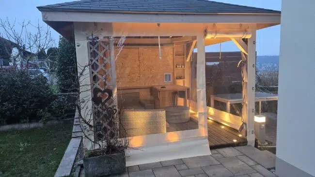 Ein gemütlicher Holzpavillon mit warmer Beleuchtung, Carport- oder Terrassenplane mit 2x Fenster und 1x Tür (mit Fenster), Sitzgelegenheiten im Freien, auf einer gefliesten Terrasse am Haus in der Abenddämmerung.