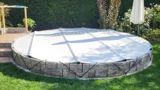 Ein runder Aufstellpool mit einer runden Poolplanen-Stangenabdeckung, die mit schwarzen Bändern befestigt ist. Umgeben von Gras, einer Steinmauer und üppigem Grün lugt rechts ein weißer Sonnenschirm hervor.
