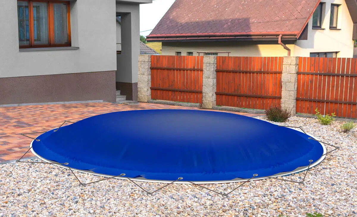 Ein blaues Trampolin mit Sicherheitsabdeckung steht auf Kies neben einem runden aufblasbaren Poolplane (Sondermaß) im Hinterhof, umgeben von einem Holzzaun und Häusern.