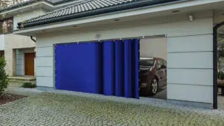 Durch ein teilweise geöffnetes Garagentor sind vor der Kulisse moderner Architektur und einer Auffahrt aus Kopfsteinpflaster ein rotes Auto und ein Schiebeplane zu sehen.