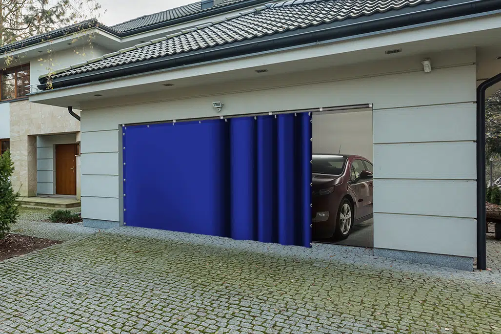 Durch ein teilweise geöffnetes Garagentor sind vor der Kulisse moderner Architektur und einer Auffahrt aus Kopfsteinpflaster ein rotes Auto und ein Schiebeplane zu sehen.