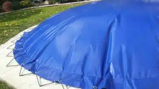 Eine große blaue Plane bedeckt ein Objekt ähnlich wie „Ovale aufblasbare Poolplanen“ auf einer kreisförmigen Betonfläche, befestigt mit Metallpflöcken. Es befindet sich auf einer grasbewachsenen Außenfläche mit Büschen und einer Holzkonstruktion dahinter.