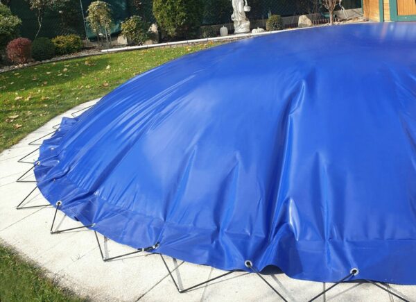 Eine große blaue Plane bedeckt ein Objekt ähnlich wie „Ovale aufblasbare Poolplanen“ auf einer kreisförmigen Betonfläche, befestigt mit Metallpflöcken. Es befindet sich auf einer grasbewachsenen Außenfläche mit Büschen und einer Holzkonstruktion dahinter.