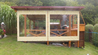 Ein hölzerner Pavillon mit Schiebeplane und Fenster ziert einen Rasenhof und bietet Stühle, einen Tisch und einen überdachten Bereich mit mehreren Gegenständen, alles inmitten von lebendigem Grün.