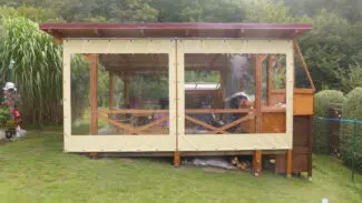 Ein hölzerner Pavillon mit Schiebeplane und Fenster ziert einen Rasenhof und bietet Stühle, einen Tisch und einen überdachten Bereich mit mehreren Gegenständen, alles inmitten von lebendigem Grün.