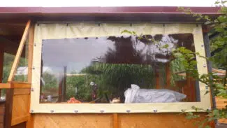 Auf einem Holzdeck befindet sich eine Schiebeplane mit Fenster, die mit Ösen befestigt ist, so dass Pflanzen draußen gedeihen können. Drinnen bleiben die Gartenmöbel unter einem braunen Metalldach geschützt.