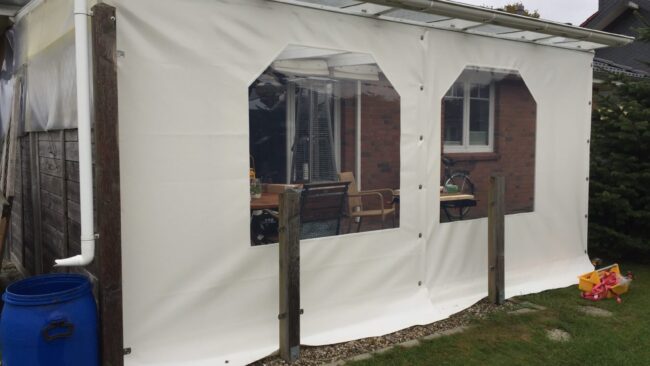 2 ttt(2) Ein weißer Terrassenplanen mit zwei durchsichtigen Kunststofffenstern bedeckt eine Terrasse, die an ein Haus angrenzt. Im Inneren sind ein Holztisch und Stühle zu sehen. Daneben steht eine blaue Regentonne und draußen auf dem Gras liegt ein rosa Spielzeug.