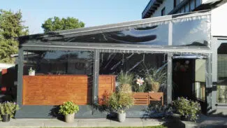 Eine moderne Terrasse mit einer transparenten „Terrassenplane in Dreiecksform mit Fenster (Links)“, Holzakzenten und Topfpflanzen, inmitten von Grün, im Hintergrund ist ein Teil eines Gebäudes sichtbar.