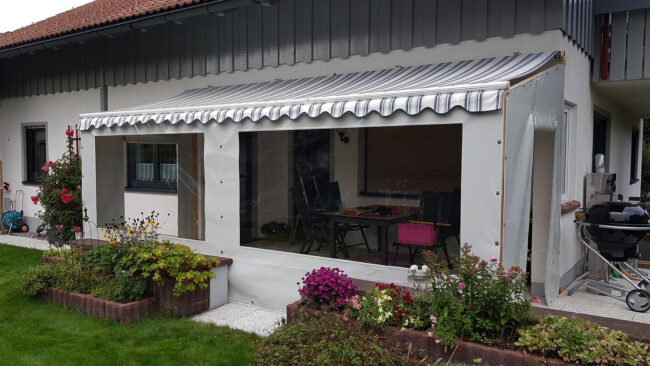 bsp1 Ein überdachter Terrassenbereich verfügt über transparente Kunststoffwände, eine gestreifte Markise und wird durch Terrassenplanen aufgewertet. Im Inneren erwarten Sie ein Tisch und Stühle inmitten farbenfroher Blumen und üppiger grüner Pflanzen im umliegenden Garten.