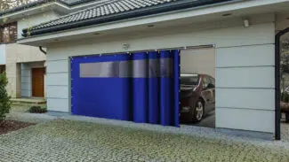 Der halb geöffnete blaue Schiebeplan mit Fenster einer modernen Garage gibt den Blick auf ein rotes Auto frei. Die grauen Wände, das Ziegeldach und der Steinbelag, der zu einer hölzernen Eingangstür führt, verleihen der Garage zusätzlichen Stil.