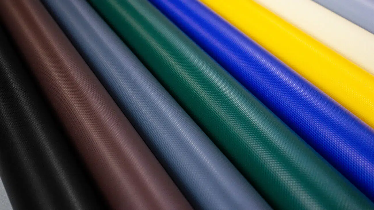 Eine Nahaufnahme von PVC-Planenrollen in verschiedenen Farben: Braun, Grau, Blau, Rot, Grün, Gelb und Beige. Diese strukturierten Stoffe haben eine matte Oberfläche und sind parallel ausgerichtet, um ein lebendiges Bild zu erzeugen.