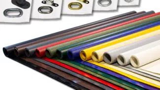 Ausgestellt sind gerollte Platten aus verschiedenfarbigen Materialien, wie LKW-Planen in Rot, Blau, Grün, Gelb und Grau. Darüber sind kleine Ösen aus Metall und Kunststoff in verschiedenen Designs auf weißem Untergrund angebracht.