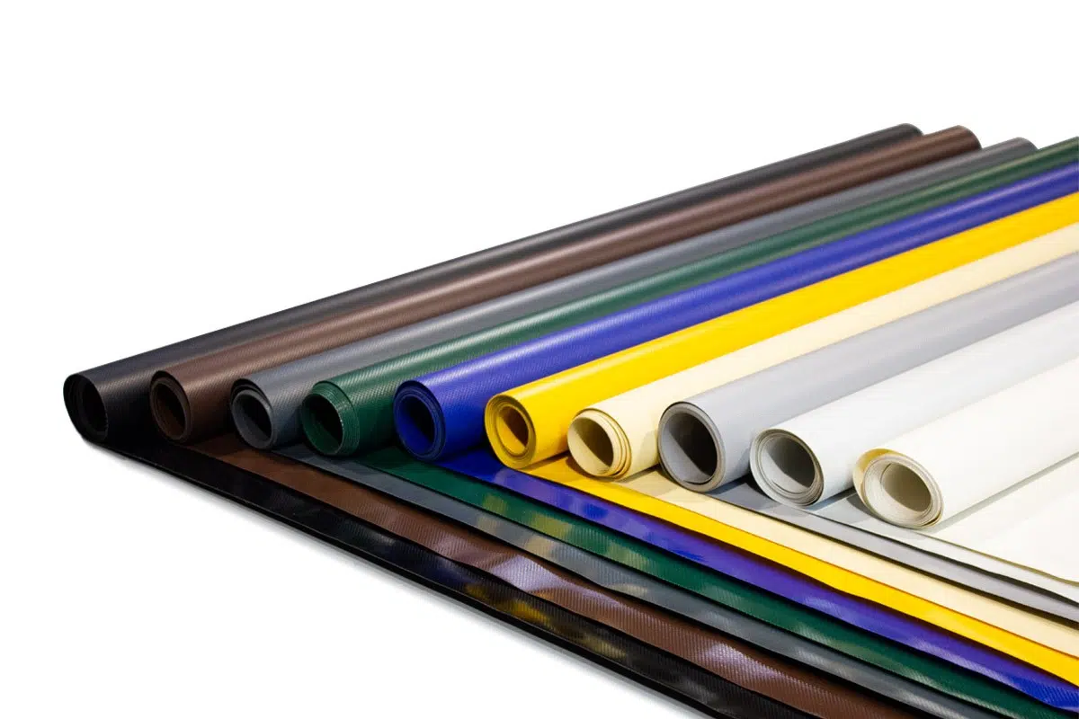 Mehrere Stoffrollen liegen in ordentlichen Reihen und zeigen eine Vielzahl von Farben. Die Stoffe umfassen Schattierungen von Schwarz, Braun, Grün, Blau, Gelb, Grau und Weiß und zeigen eine Mischung aus Texturen und Verarbeitungen.