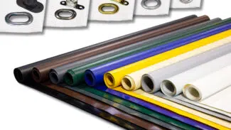 Rollen mit buntem Stoff für LKW Planen sind in einer mehrschichtigen Präsentation angeordnet und weisen Farbtöne wie Schwarz, Grün und mehr auf. Darüber sind verschiedene Ösen aus Metall und Kunststoff ausgelegt, die unterschiedliche Designs zeigen.