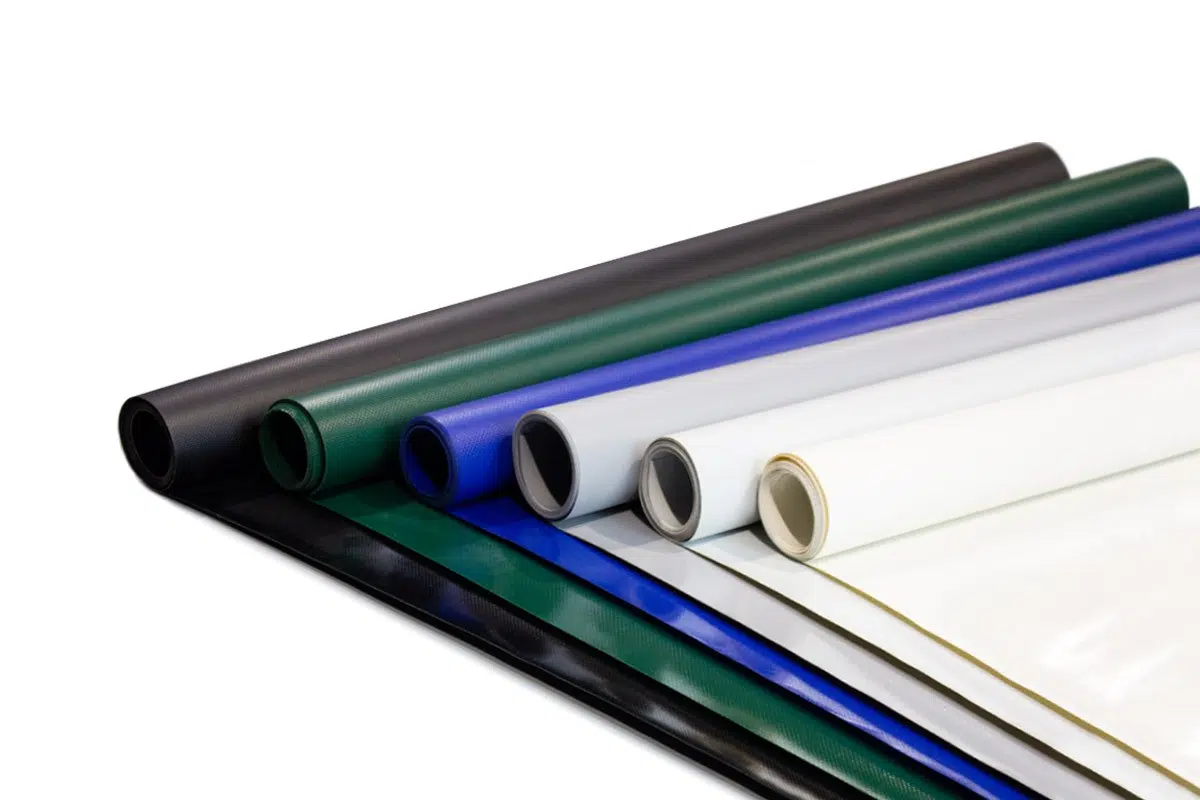 Ein Satz aufgerollter Vinylfolien, die an LKW Planen erinnern, in Farben wie Schwarz, Grün, Blau, Weiß und Creme. Ordentlich auf einer weißen Oberfläche angeordnet, zeigen sie unterschiedliche Texturen und Oberflächen.