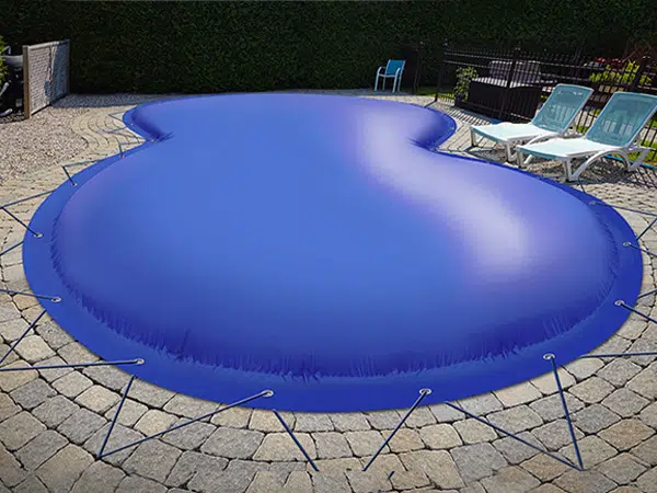 Ein Pool im Hinterhof, der mit einer festsitzenden, aufgeblasenen blauen Poolplane-Abdeckung abgedeckt ist, erzeugt einen blasenartigen Effekt. Auf der Steinterrasse in der Nähe stehen zwei leere Liegestühle.
.
