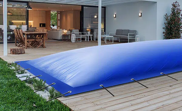 Eine moderne Terrasse mit Holzdeck bietet Sitzgelegenheiten im Freien und einen Essbereich. Eine große blaue Poolplane bedeckt elegant einen Teil des Decks und des Rasens, während Glastüren den Blick auf das stilvolle Innere des Hauses freigeben.