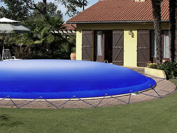 Eine große, blaue Poolplane-Kuppel überdeckt den Außenpool, gesichert mit Seilen. Im Hintergrund sind unter einem leicht bewölkten Himmel ein Haus mit Holzläden und ein Garten mit Bäumen zu sehen.