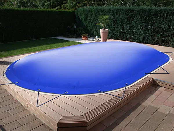 Ein Pool im Hinterhof ist mit einer blauen, aufblasbaren Poolplane abgedeckt, die sicher am Holzdeck befestigt ist. Der Garten verfügt über sorgfältig angelegte Hecken und farbenfrohe Topfpflanzen.