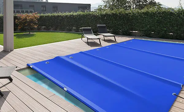 Ein rechteckiger Swimmingpool, der mit einer hellblauen Poolplane bedeckt ist, ist von einem Holzdeck umgeben. Auf dem Deck stehen zwei schwarze Sonnenliegen mit kleinen Tischen, während üppige grüne Hecken und Bäume eine ruhige Kulisse bilden.