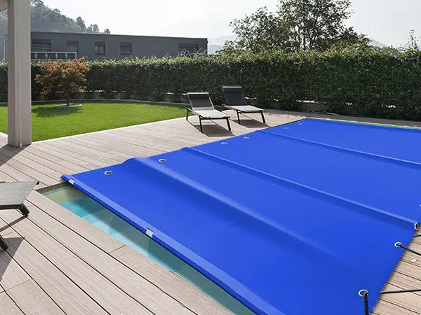 Ein rechteckiger Swimmingpool, der mit einer hellblauen Poolplane bedeckt ist, ist von einem Holzdeck umgeben. Auf dem Deck stehen zwei schwarze Sonnenliegen mit kleinen Tischen, während üppige grüne Hecken und Bäume eine ruhige Kulisse bilden.