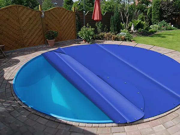 Ein kleiner, runder Pool im Hinterhof verfügt über eine blaue Poolplane-Abdeckung. Umgeben von einer gemauerten Terrasse und einem Holzzaun ist der Bereich üppig grün und verfügt über einen roten Sonnenschirm.