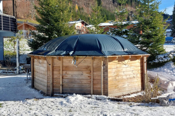 Eine runde Holzkonstruktion mit schwarzem Dach im Schnee, teilweise bedeckt von Achteckigen aufblasbaren Poolplanen.