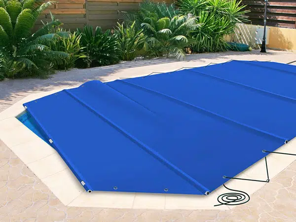 Eine blaue Poolplane ist sicher über einem Swimmingpool auf einer gefliesten Terrasse befestigt. Der Bereich ist von üppigen grünen Pflanzen und einem Holzzaun umgeben, die eine tropische Atmosphäre schaffen.