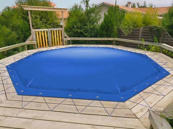 Eine blaue achteckige Poolplane deckt den Aufstellpool auf einem Holzdeck ab. Mit Kordeln und Ösen fest befestigt, steht sie zwischen Bäumen und einem Haus im Hintergrund.
