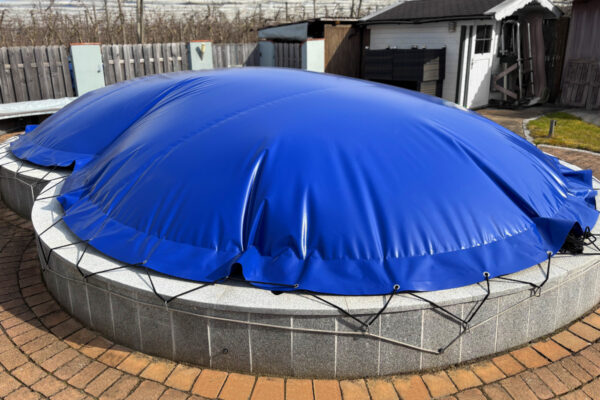 Ein großer, runder Pool ist mit den Achteckigen aufblasbaren Poolplanen abgedeckt, mit Seilen gesichert und auf einer gemauerten Terrasse in einem eingezäunten Hof aufgestellt.