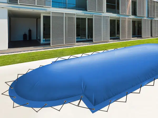 Ein großes blaues Poolplane ist mit schwarzen Gurten sicher über einem Außenpool befestigt. Neben einem modernen Gebäude blicken Terrassentüren und Lamellenpaneele auf das üppige grüne Gras, das die Umgebung umgibt.