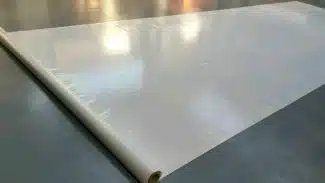 Eine große Rolle weißen, glänzenden Materials, teilweise ausgerollt auf einer glatten, reflektierenden Oberfläche. Der Glanz des Materials erinnert an eine Vinyl- oder Kunststoffzusammensetzung und das Sonnenlicht strömt sanft über die Oberfläche.