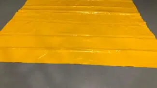 Auf dem grauen Betonboden liegt eine große, flache gelbe LKW PVC Planen Reste Plane. Ihre glänzende Oberfläche und die sichtbaren Falten fangen das Licht wunderschön ein.
