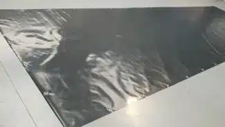 Eine große, glänzende, dunkelgraue Plane auf einem glatten, hellen Boden ähnelt LKW PVC Planen Reste. An den Rändern befinden sich Metallösen, die das Licht reflektieren und eine leicht faltige Textur aufweisen.