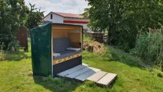 Eine kleine grüne Abdeckhaube vom Typ Sonderform 1 mit offener Vorderseite und Bank steht auf einer Rasenfläche, umgeben von Bäumen und Grünflächen. Im Hintergrund lugt ein Gebäude mit rotem Dach hervor.