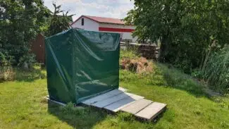 Auf einer von Bäumen und Sträuchern umgebenen Rasenfläche steht ein begrünter Baukörper mit Schrägdach, ähnlich den Abdeckhauben Sonderform 1. Ein mit Betonplatten ausgelegter Weg führt dorthin, im Hintergrund ist ein Gebäude zu erkennen.