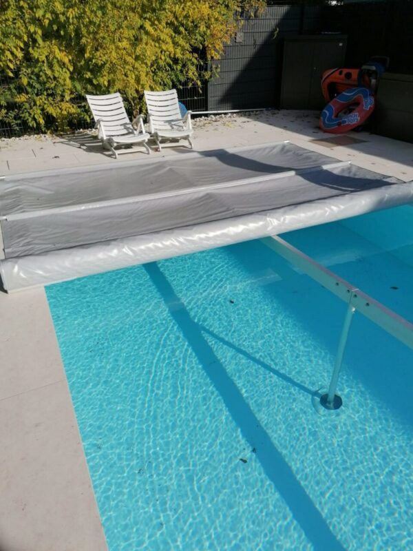 Ein Swimmingpool mit teilweise ausgefahrener silberner Abdeckung, zwei weißen Liegestühlen daneben und einem leuchtend orangefarbenen Schwimmkörper vor einem Zaun mit grünem Laub. Kaufen Sie noch heute Schneeträger für Stangenabdeckungen!.