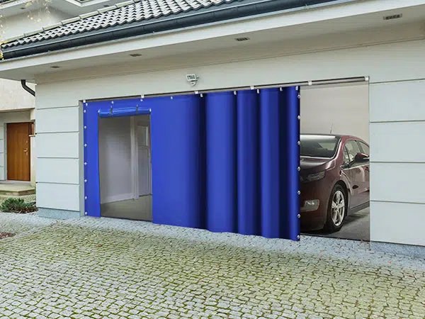 Eine Garage mit einem teilweise geöffneten blauen Schiebeplanen gibt den Blick auf ein darin geparktes Auto frei. Die Garage, die an ein helles Haus mit Ziegeldach angebaut ist, verfügt über eine mit Kopfstein gepflasterte Auffahrt.