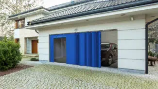 Ein modernes Haus mit geschlossener Holztür und teilweise geöffneter blauer Schiebetür zur Garage, hinter der ein geparktes Auto zu sehen ist. Die Garage hat eine gepflasterte Auffahrt, die von Grün umgeben ist.