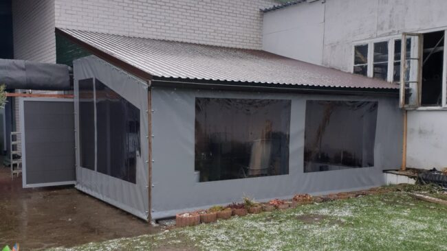 Terrassenplane Neben einem Backsteingebäude steht ein kleines Vordach mit schrägem Metalldach und Kunststofffenstern. Am Rand stehen schneebedeckte Terrassenplanen, im Inneren sind Pflanzen zu sehen, die auf die wärmeren Monate hinweisen.