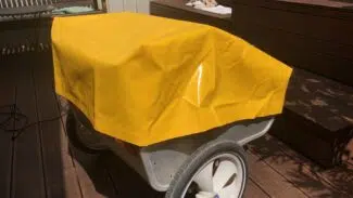 Ein kleiner Wagen mit großen Rädern steht auf einem Holzdeck und ist mit einer leuchtend gelben Plane bedeckt, die einer Abdeckhaube Sonderform 8 ähnelt. Im Hintergrund sind eine Bank und Stufen zu sehen, auf denen Schuhe zu sehen sind.