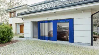 Ein modernes Haus mit weißer Außenfassade und grauem Ziegeldach hat ein teilweise geöffnetes Garagentor, durch das ein Auto zu sehen ist. Die Einfahrt mit Steinfliesen kontrastiert mit dem Grün und einer Schiebeplane mit Fenster/mit Tür an der Seite.