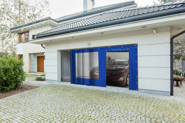Ein modernes Haus mit weißer Außenfassade und grauem Ziegeldach hat ein teilweise geöffnetes Garagentor, durch das ein Auto zu sehen ist. Die Einfahrt mit Steinfliesen kontrastiert mit dem Grün und einer Schiebeplane mit Fenster/mit Tür an der Seite.