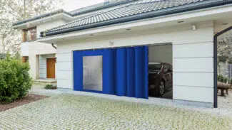 Ein Haus hat eine Garage mit einem blauen Vorhang wie die Schiebeplane mit Fenster/mit Tür, der den Eingang teilweise abdeckt. Im Inneren steht ein geparktes Auto, während die gepflasterte Auffahrt und das Grün einen ruhigen Vordergrund schaffen.