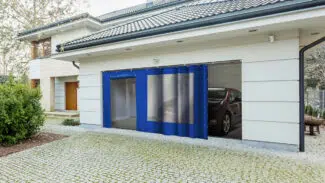 Modernes Haus mit sichtbarer Garage mit Schiebeplane mit Fenster/mit Tür. Im Inneren ist die Vorderseite eines Autos geparkt. Die Außenseite hat ein Ziegeldach, eine gepflasterte Auffahrt und Büsche auf der linken Seite.