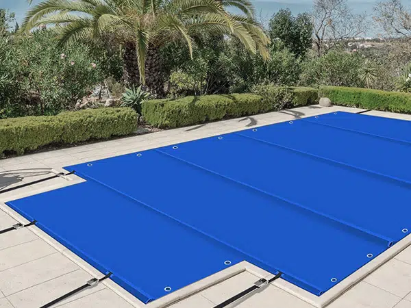 Ein Swimmingpool mit einer leuchtend blauen Poolplane-Abdeckung akzentuiert die Steinterrasse, flankiert von üppigen grünen Sträuchern und einer hoch aufragenden Palme unter dem klaren blauen Himmel.