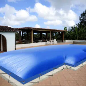 In einem geräumigen Innenhof befindet sich ein Swimmingpool, der mit einer straff gespannten blauen Abdeckung bedeckt ist. Der Bereich ist von Grün umgeben und verfügt über einen überdachten Sitzbereich im Freien mit Holzmöbeln. Am blauen Himmel darüber sind flauschige Wolken zu sehen.