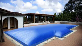 In einem geräumigen Innenhof befindet sich ein Swimmingpool, der mit einer straff gespannten blauen Abdeckung bedeckt ist. Der Bereich ist von Grün umgeben und verfügt über einen überdachten Sitzbereich im Freien mit Holzmöbeln. Am blauen Himmel darüber sind flauschige Wolken zu sehen.