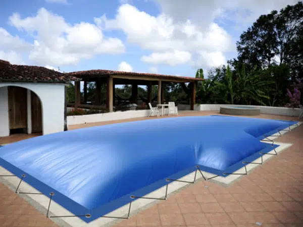 Ein Hinterhof mit einem mit Poolplane abgedeckten Pool, der mit Gummiseilen und Haken gesichert ist. Er ist umgeben von einem gefliesten Deck, Bäumen und einem gemütlichen Sitzbereich unter einem Baldachin, alles unter einem teilweise bewölkten Himmel.